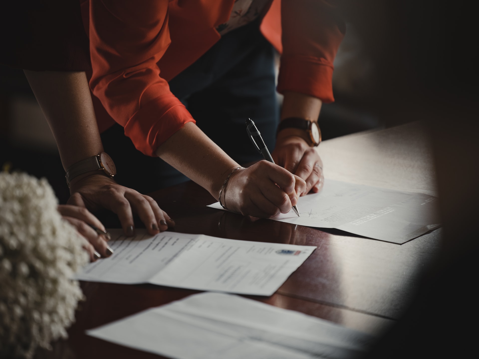 Person in an orange blazer signing a document on a wood table.