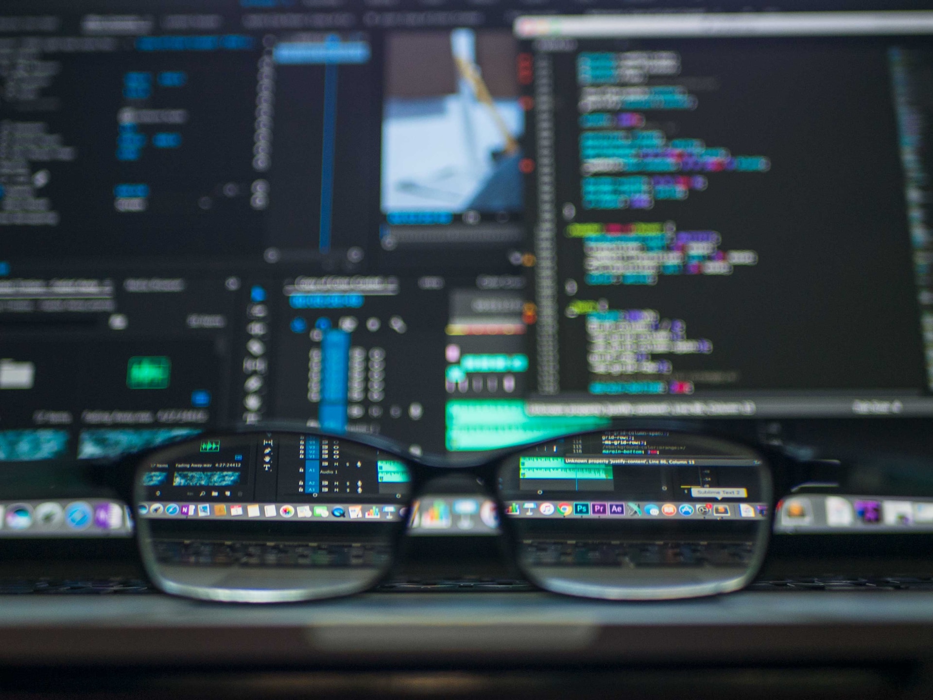 Eyeglasses resting on a desk with computer monitors in the background